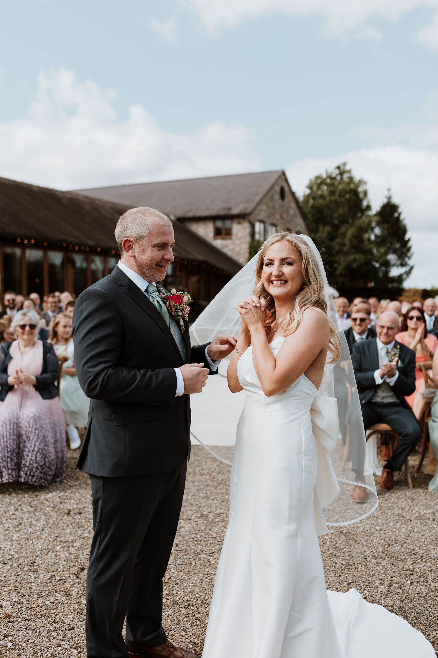 A couple celebrates during an outdoor wedding ceremony with guests seated on patio seating behind rustic barn buildings.