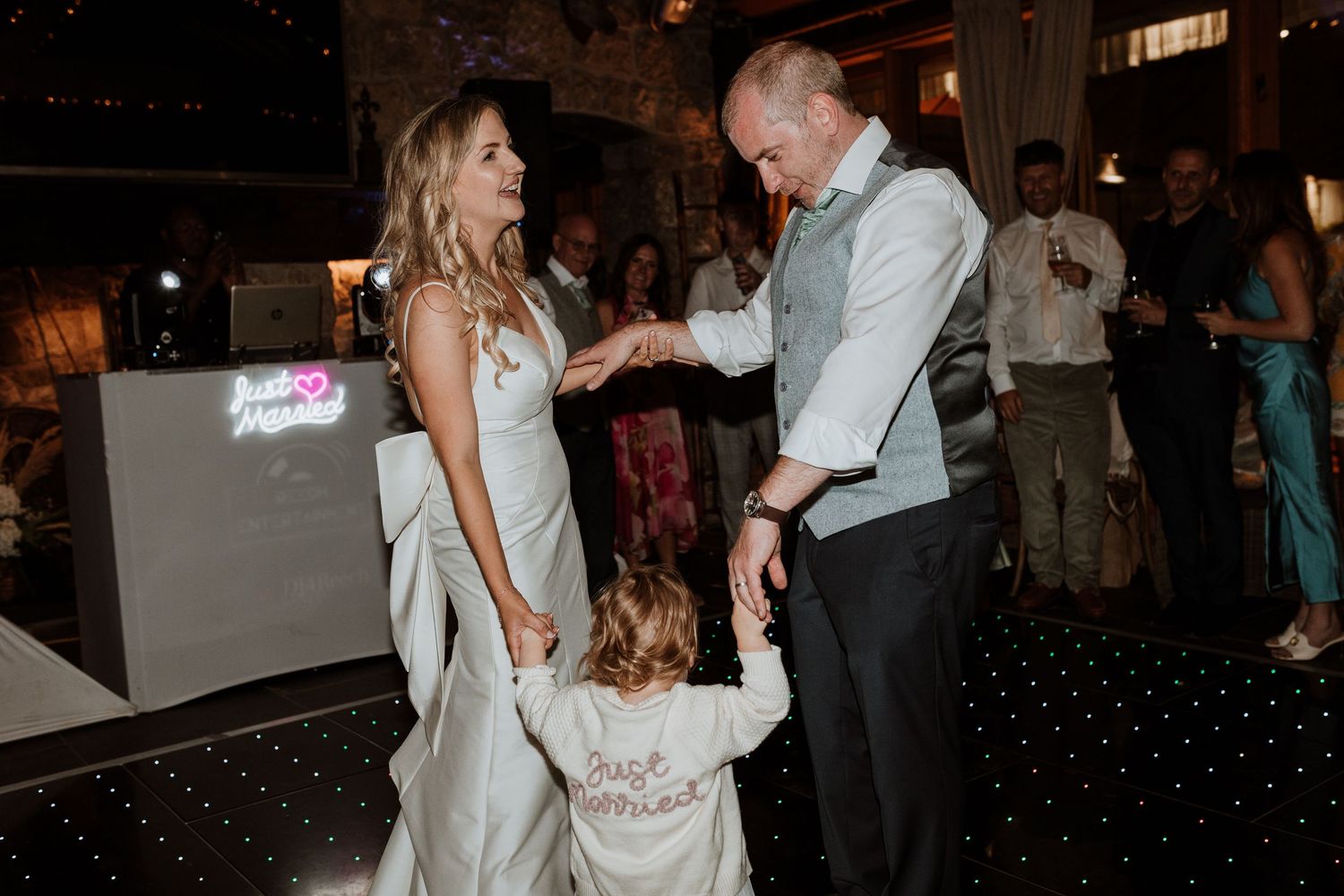 A newlywed couple shares a special dance moment with a young child on an illuminated dance floor at their wedding reception.