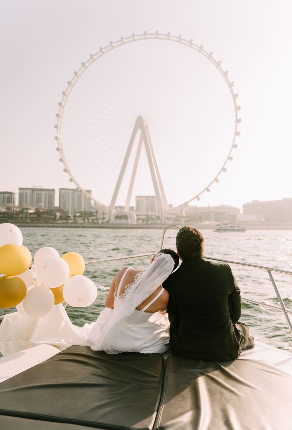 Couple sits on a boat with balloons while viewing the Dubai Eye observation wheel over the water at sunset.