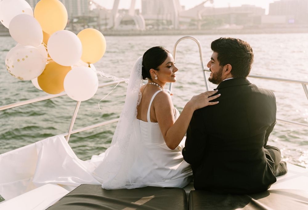 Romantic couple sitting on a boat with white and yellow balloons against a city waterfront backdrop.