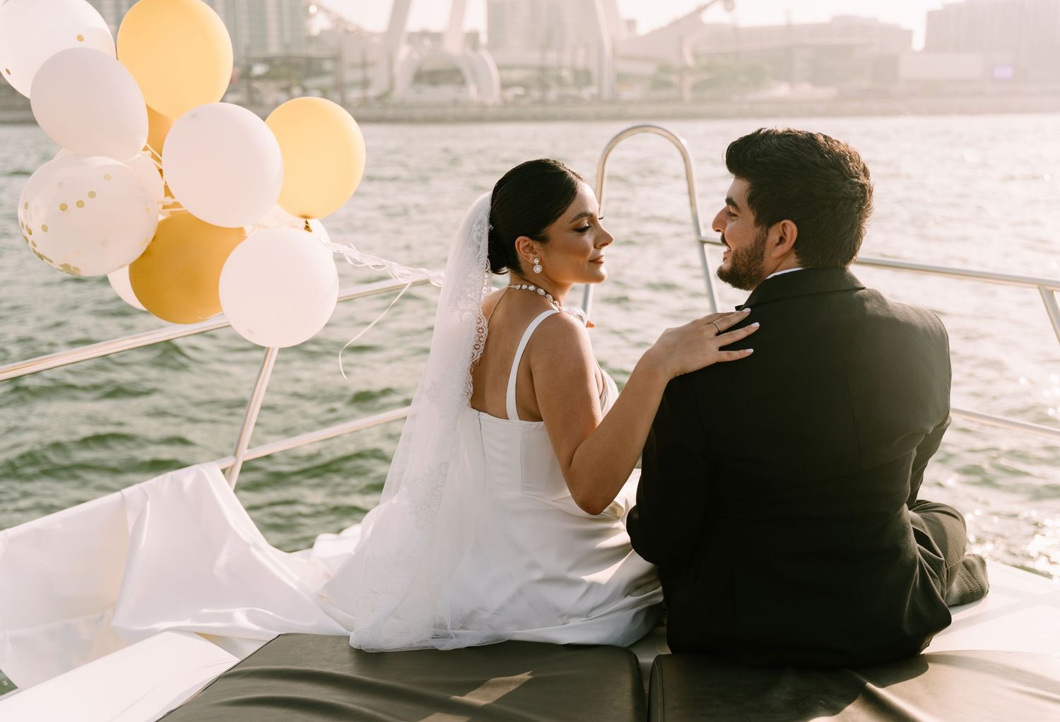 Romantic couple sitting on a boat with white and yellow balloons against a city waterfront backdrop.