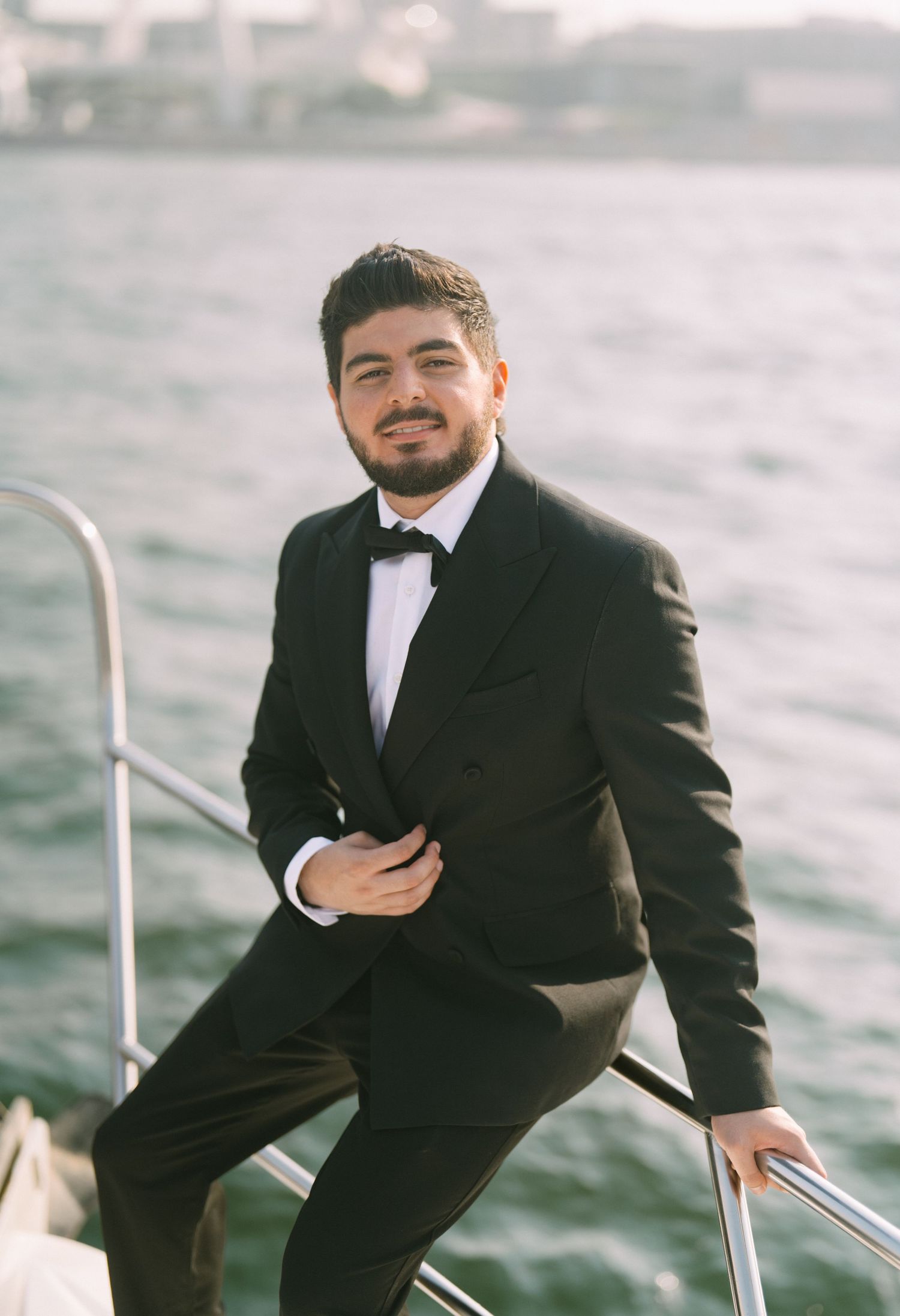 A person in a black tuxedo and bow tie stands on a boat deck with water in the background during daytime.