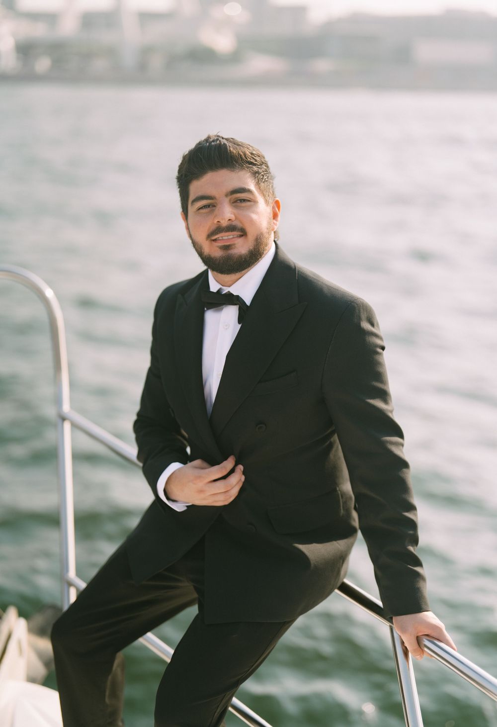 A person in a black tuxedo and bow tie stands on a boat deck with water in the background during daytime.