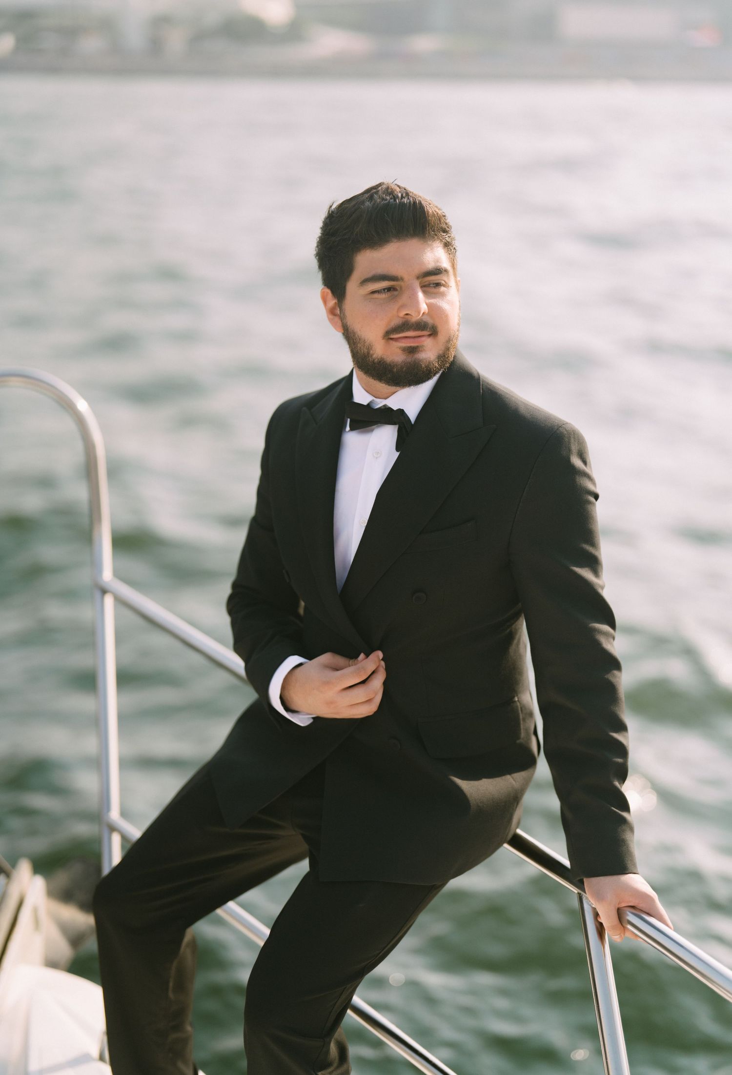 A gentleman in a black tuxedo sits casually on the railing of a boat while on the water.
