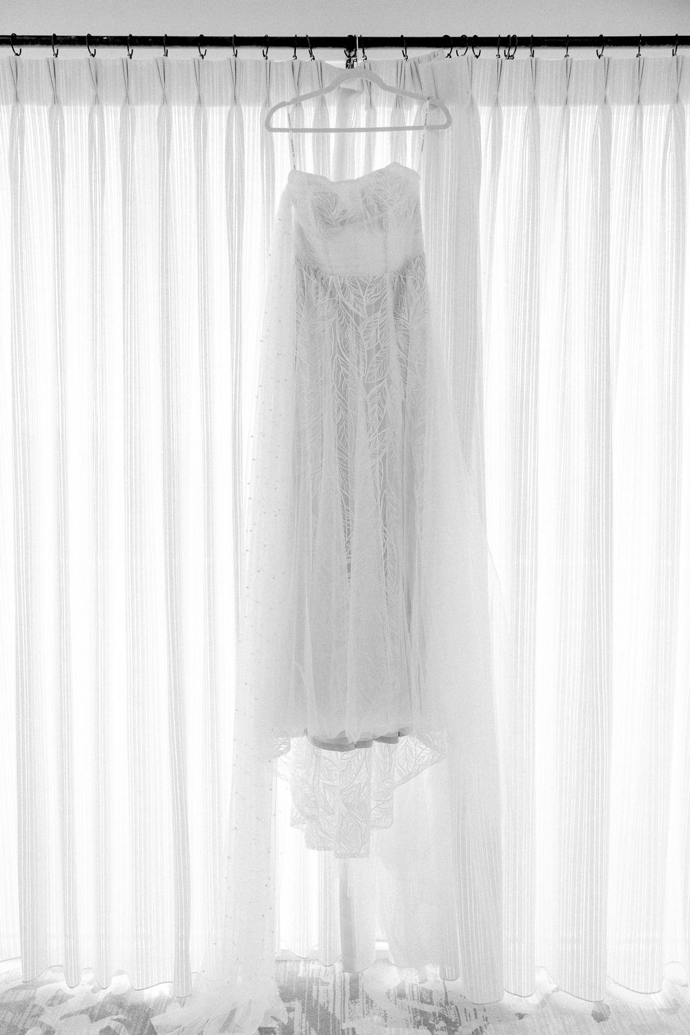 A white wedding dress hangs elegantly in front of sheer curtains in black and white photography.