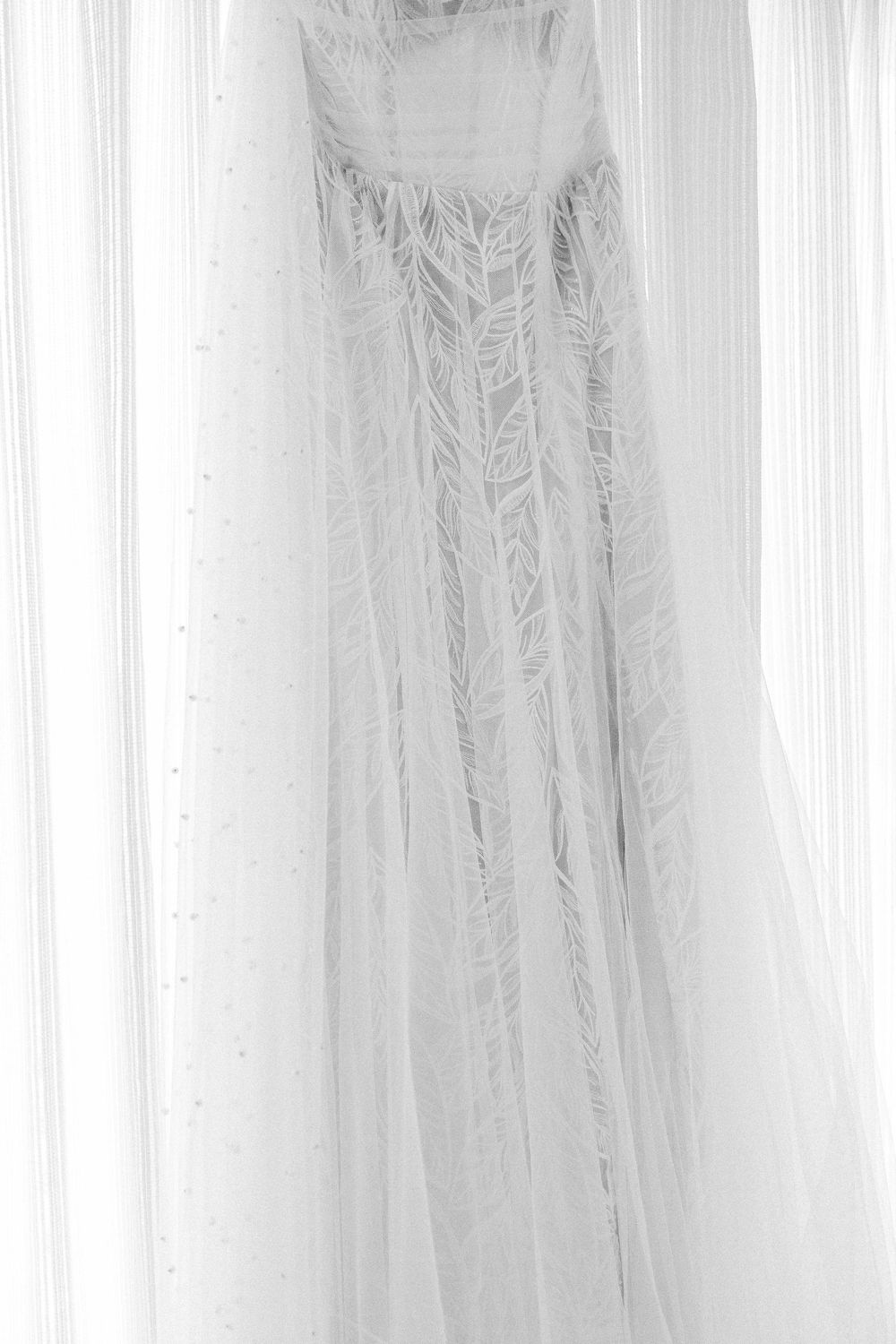 A delicate white wedding dress with intricate lace detail hangs elegantly against a light background.