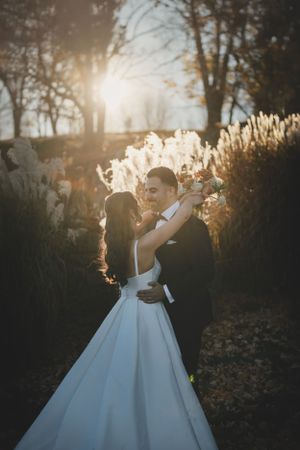 A newlywed couple embraces as golden sunlight filters through trees and tall grasses at sunset.