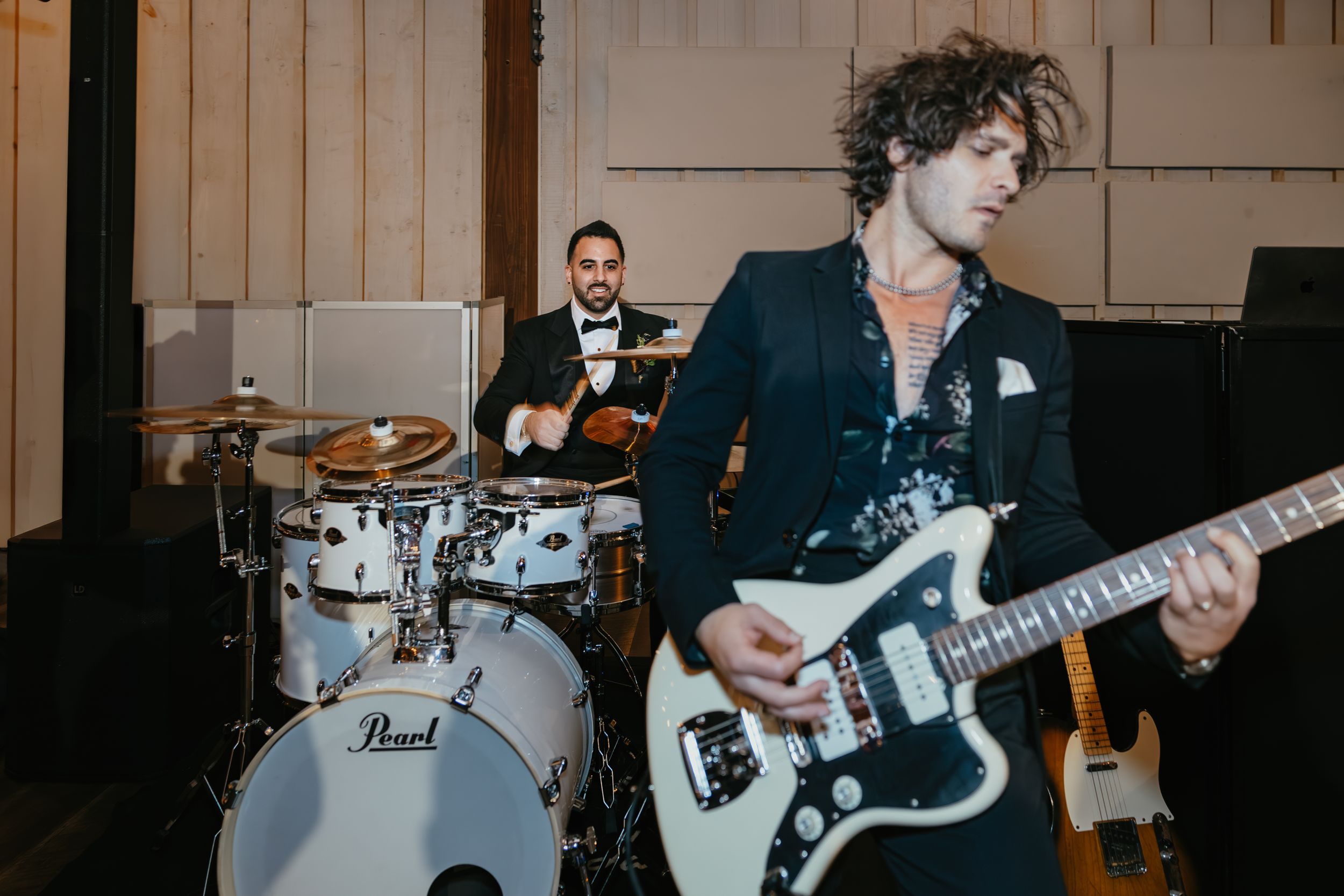 Musicians perform energetically at a venue with a drummer behind a pearl kit and guitarist playing a white bass guitar.
