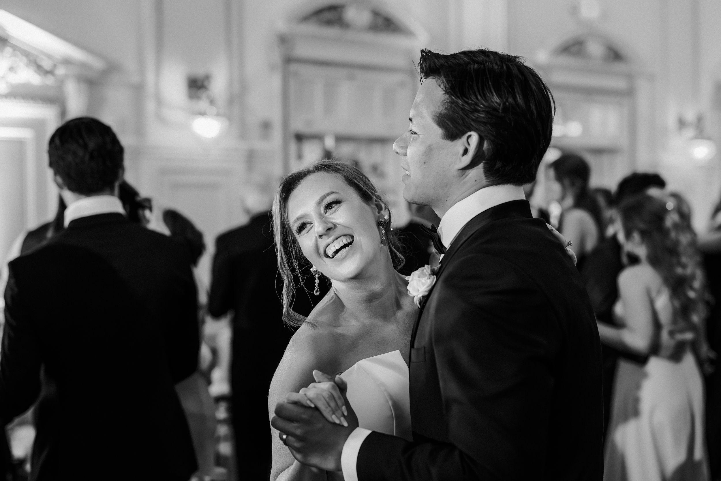 A couple shares a joyful laugh while dancing together at an elegant wedding reception in black and white.
