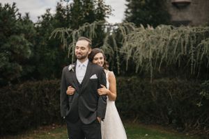 A couple stands together in formal attire against a backdrop of lush greenery and ornamental grasses at an outdoor venue.