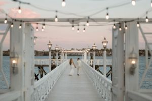 A romantic pier wedding ceremony setup with string lights and white railings during sunset over the waterfront.
