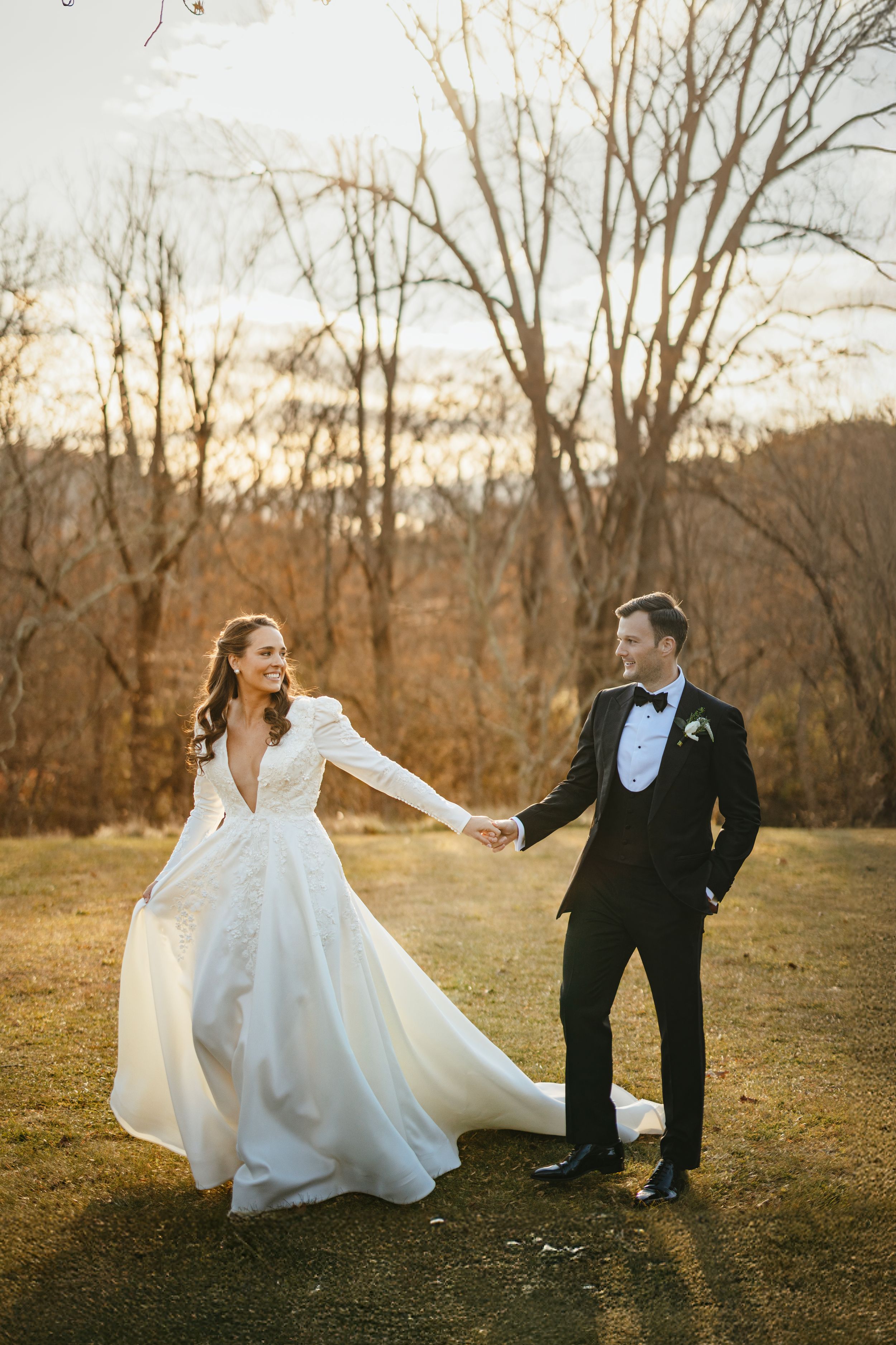 A joyful wedding couple dances together in an autumn field with bare trees and golden sunlight at sunset.