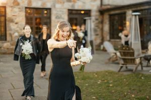 Someone in an elegant black formal dress walks outdoors while holding flowers at a stone building venue.