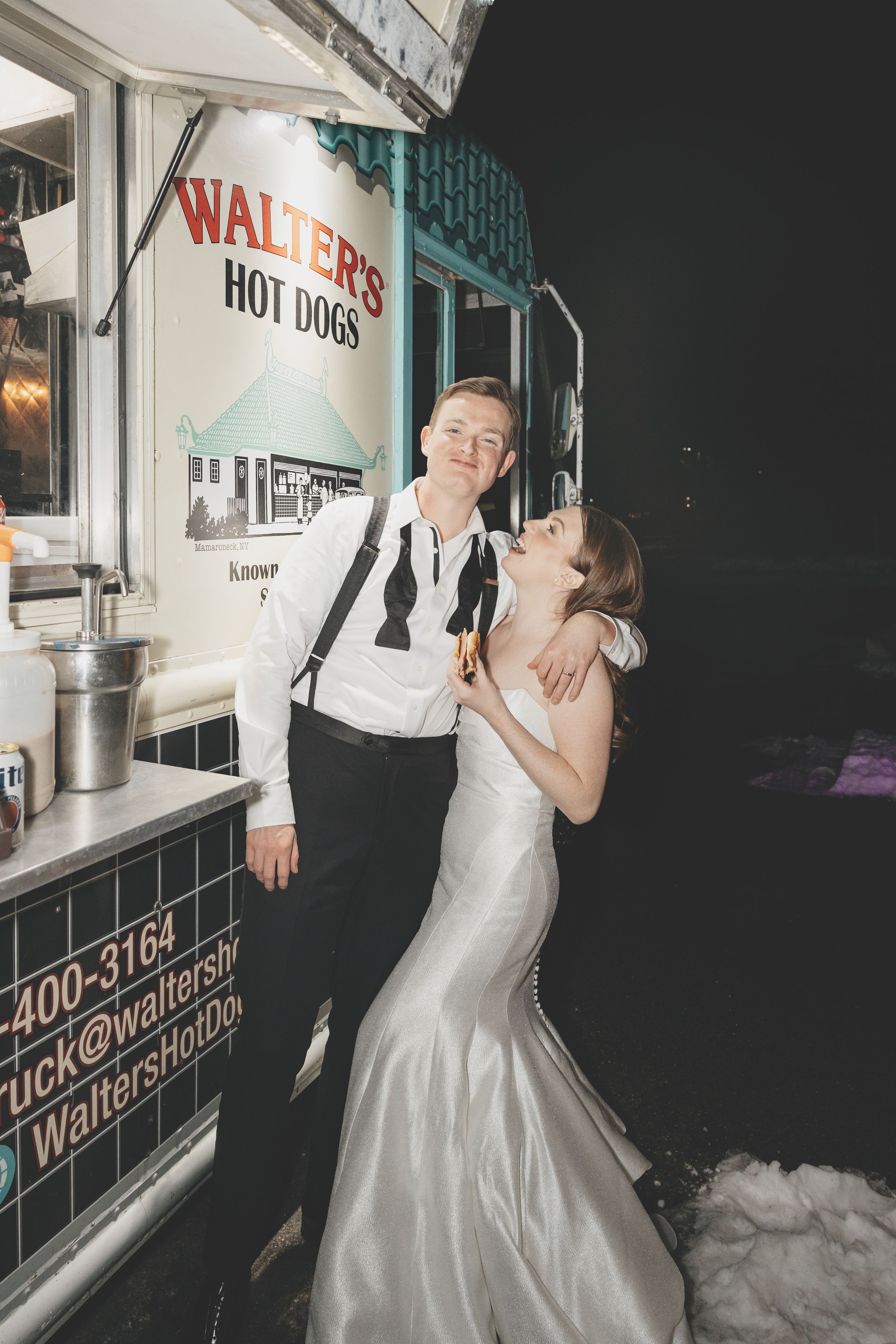Couple in formal attire poses by a hot dog stand called Walter's at nighttime in winter with snow visible on the ground.