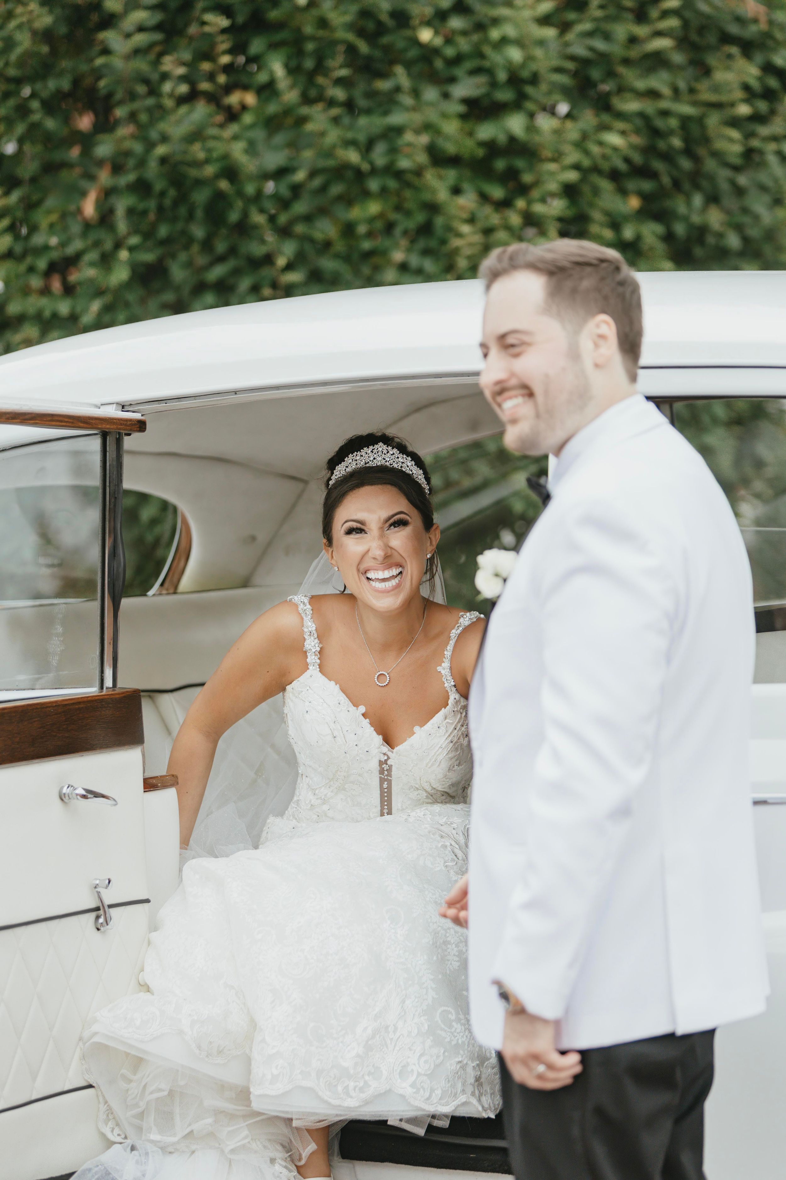 A bride and groom share a joyful moment beside a white vintage car during their wedding celebration.