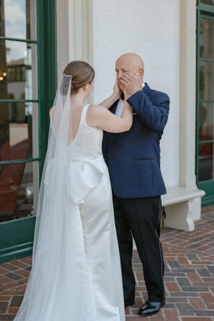 Emotional first look moment between a bride in white gown and person in navy suit on brick sidewalk.