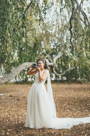 A bride in a flowing white gown poses under graceful willow tree branches on autumn ground.
