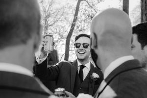 A person in sunglasses and a suit laughs while surrounded by other guests at an outdoor wedding celebration.