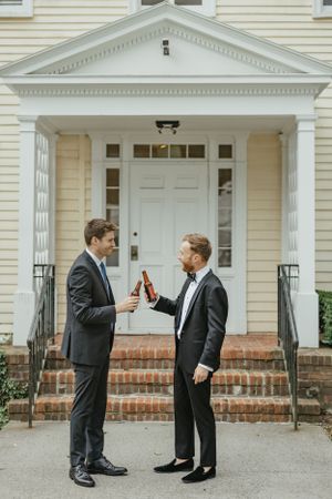 Two people in formal suits toast with drinks outside a white colonial-style building entrance with brick steps.