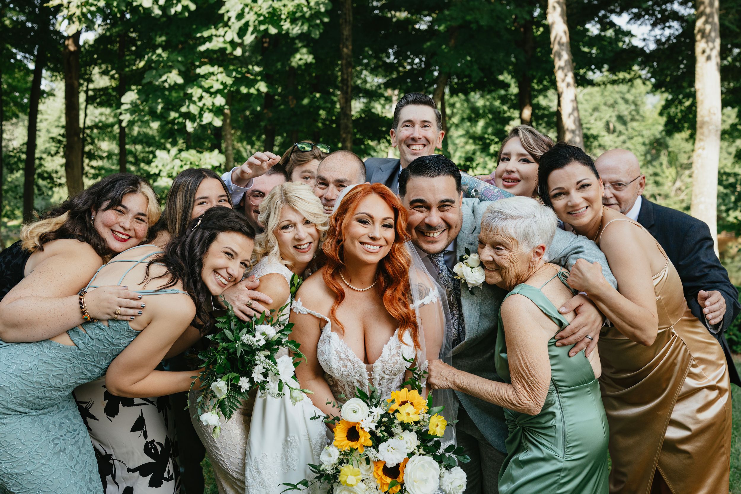 Wedding party in mix of sage green dresses huddles close together with sunflower bouquet for cheerful outdoor photo.