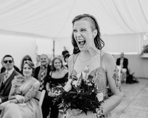 A tattooed and joyfully shouting wedding guest with dark hair holding a bouquet at an outdoor ceremony.
