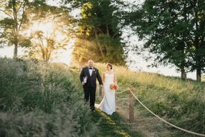 A couple walks along a grassy hillside path at golden hour with sunlight streaming through trees in the background.
