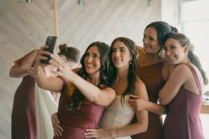 Group wearing burgundy bridesmaid dresses gather around the bride in white for a joyful selfie moment.