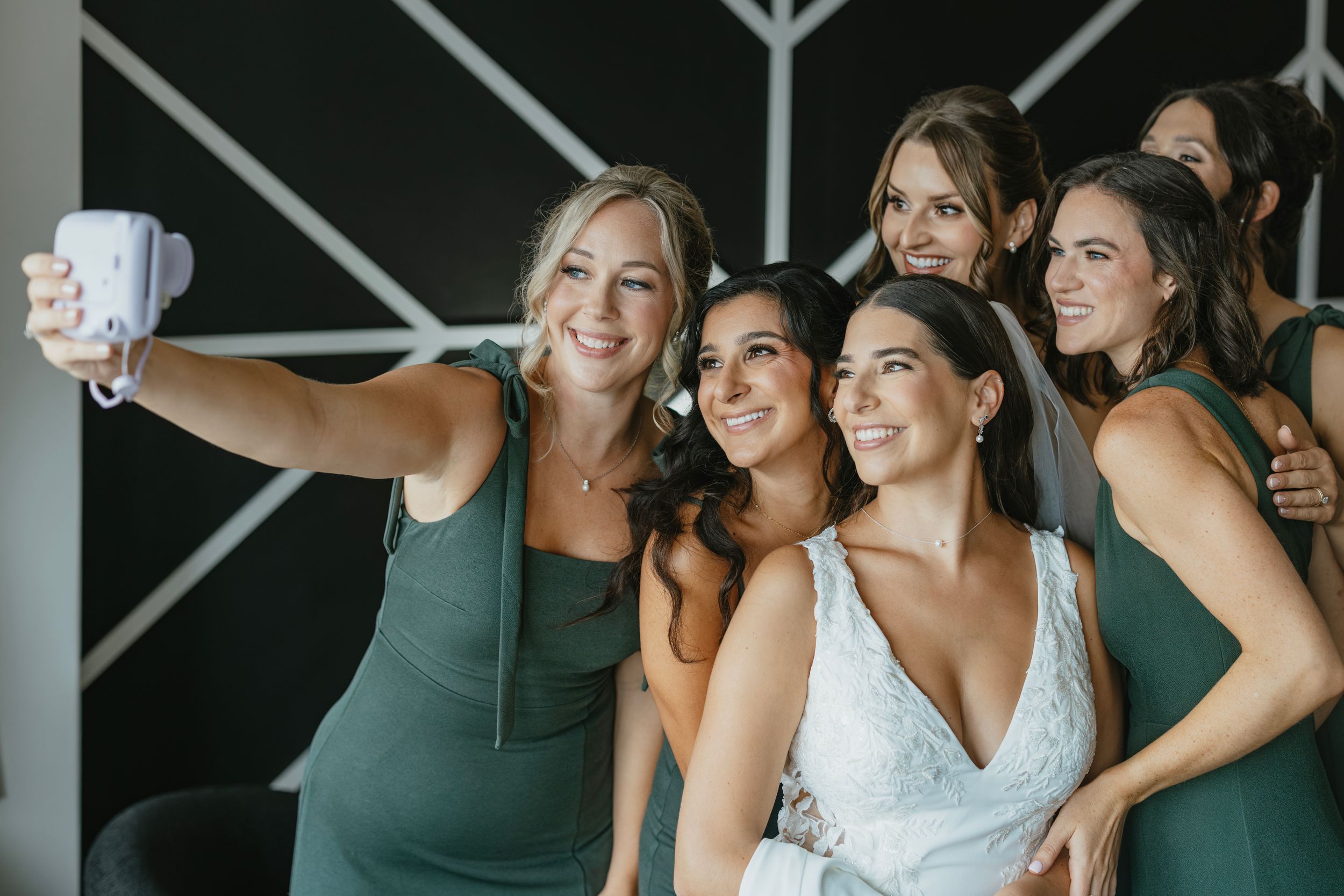 Group in matching green bridesmaid dresses taking selfies with bride in white wedding gown against geometric wall decor.