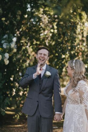 Joyful bride and groom share a candid laughing moment together outdoors with bokeh light and greenery in background.
