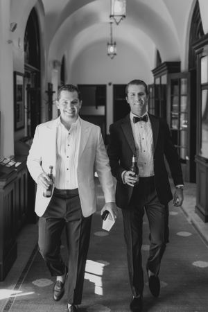 Two people in formal tuxedos walking down an arched hallway while holding beverages at a black tie event.