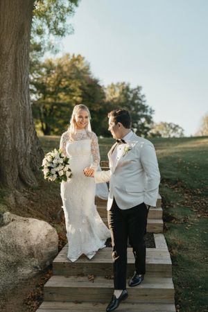 A couple in formal wedding attire stand together on wooden steps outdoors during golden hour near a large tree.