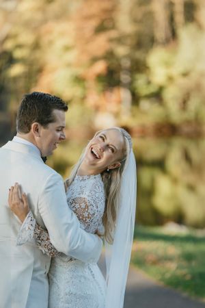 Joyful couple shares a candid laughing moment on their wedding day in white attire against autumn foliage.