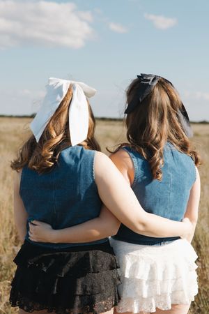 Friends in matching denim embracing in sunlit field wearing white bow and sunglasses.