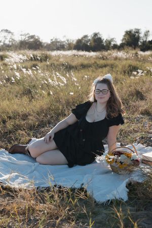 Person in a black dress lounging on white blanket in grassy field at sunset.