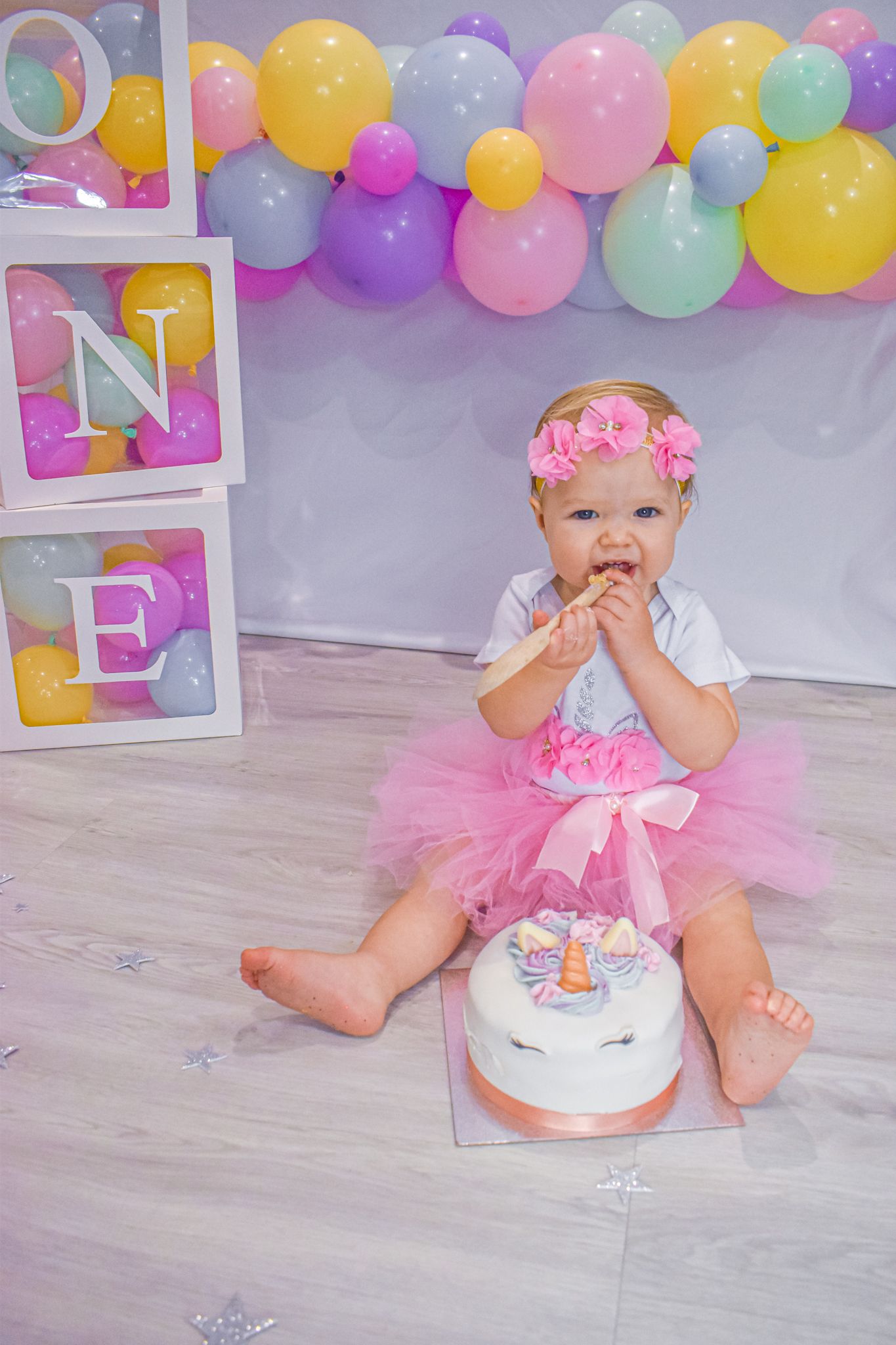 Baby in pink tutu sits with birthday cake surrounded by colorful balloons.