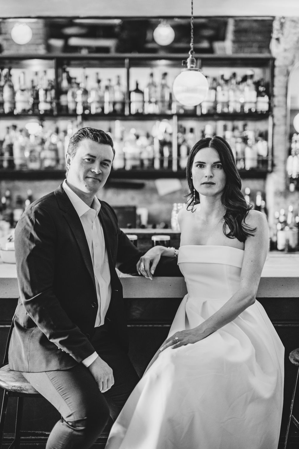 Elegant couple in wedding attire pose at stylish bar with illuminated bottles in background.