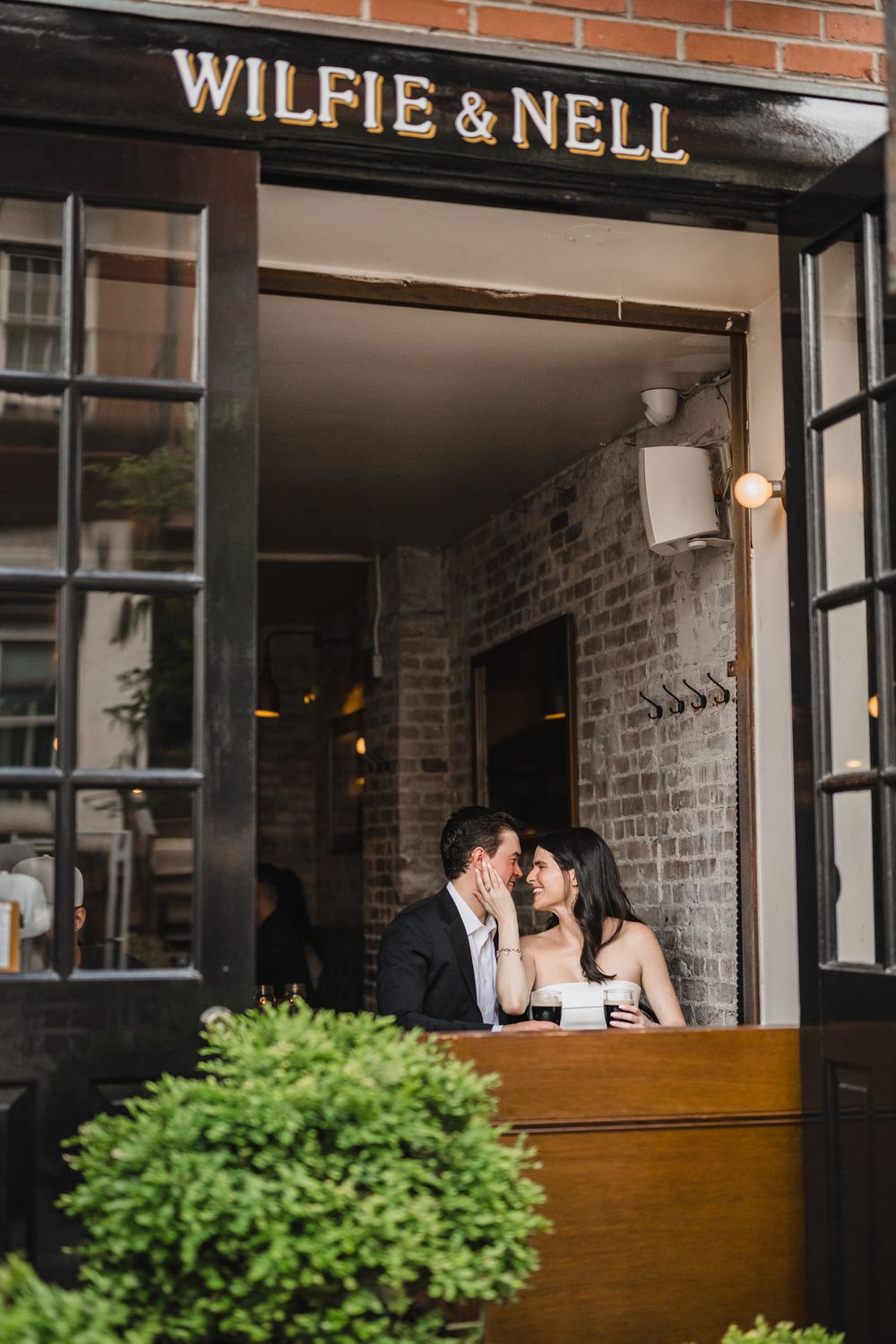 Couple shares romantic moment at Wilfie & Nell restaurant entrance with brick wall backdrop.