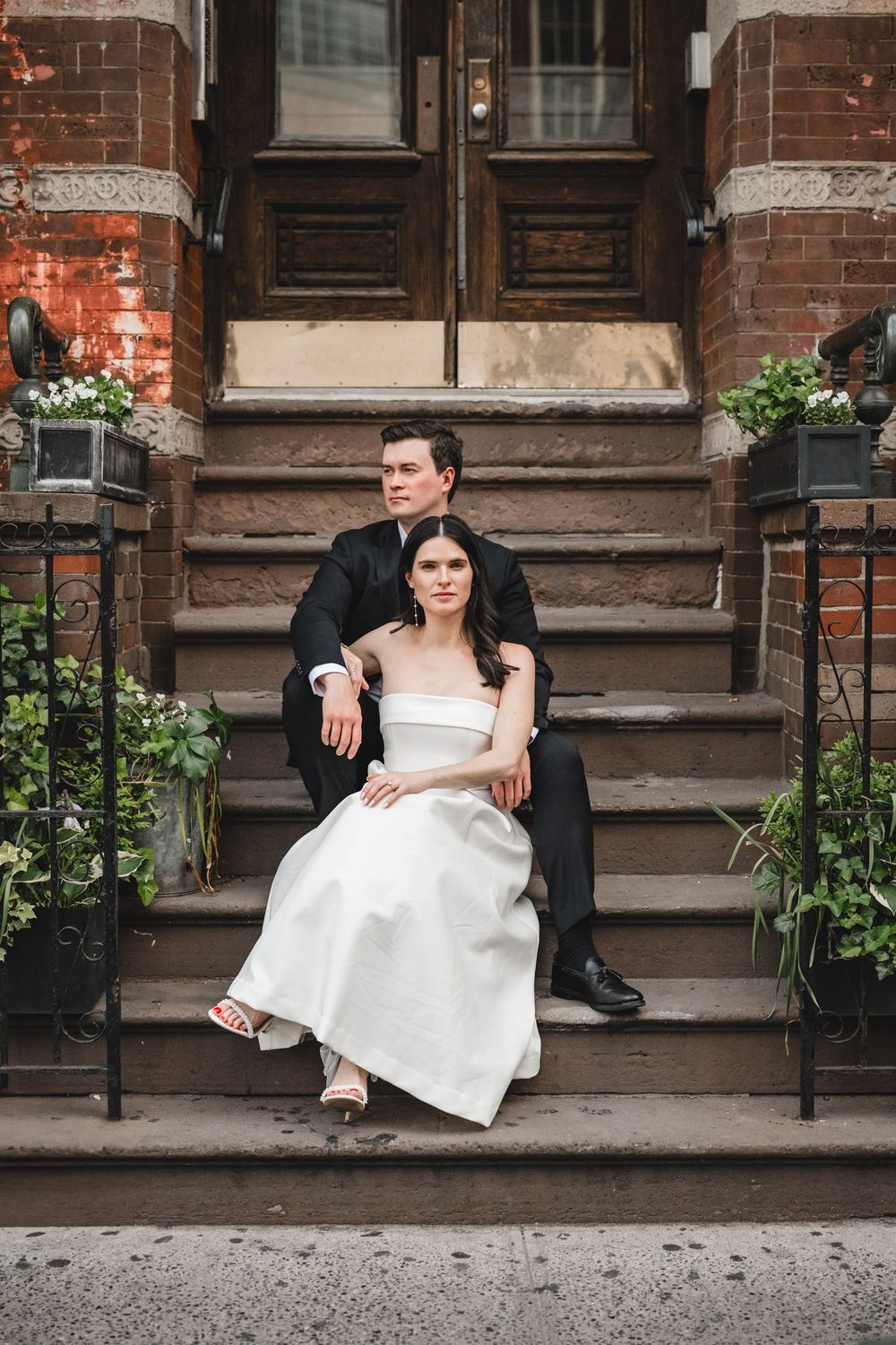 Couple in formal attire poses on brownstone steps with decorative lanterns and planters.