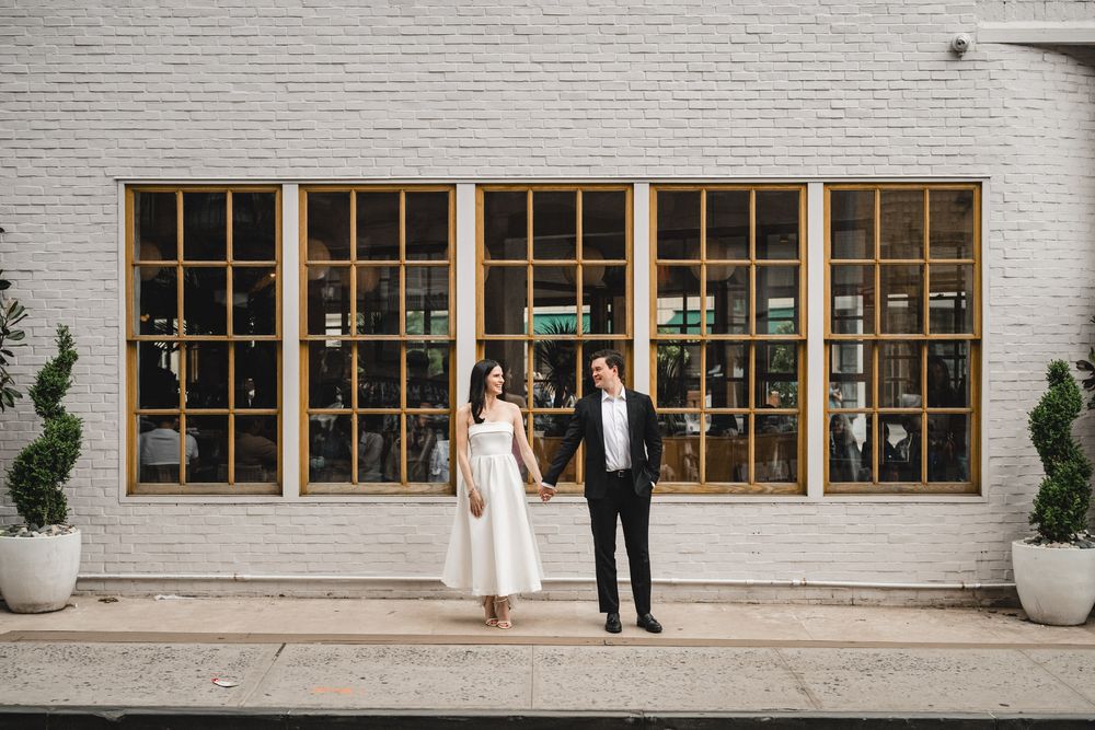 Couple in wedding attire poses against white building with window grid and potted plants.
