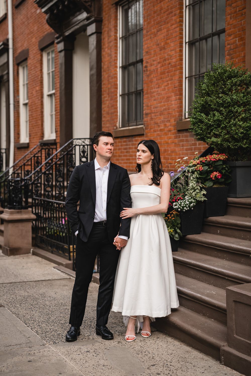 Couple in wedding attire poses on brownstone steps with red brick facade and iron railings.