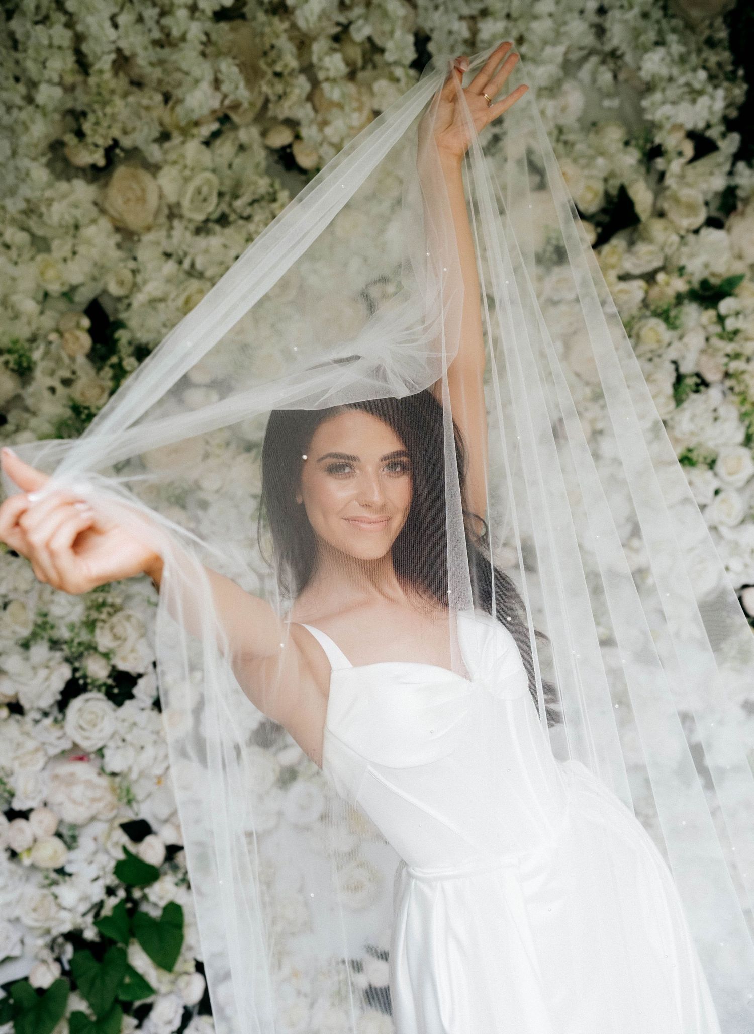 A bride in a flowing white veil poses among a romantic floral arch of white blossoms and greenery. Wedding Photographer Wedding Videographer