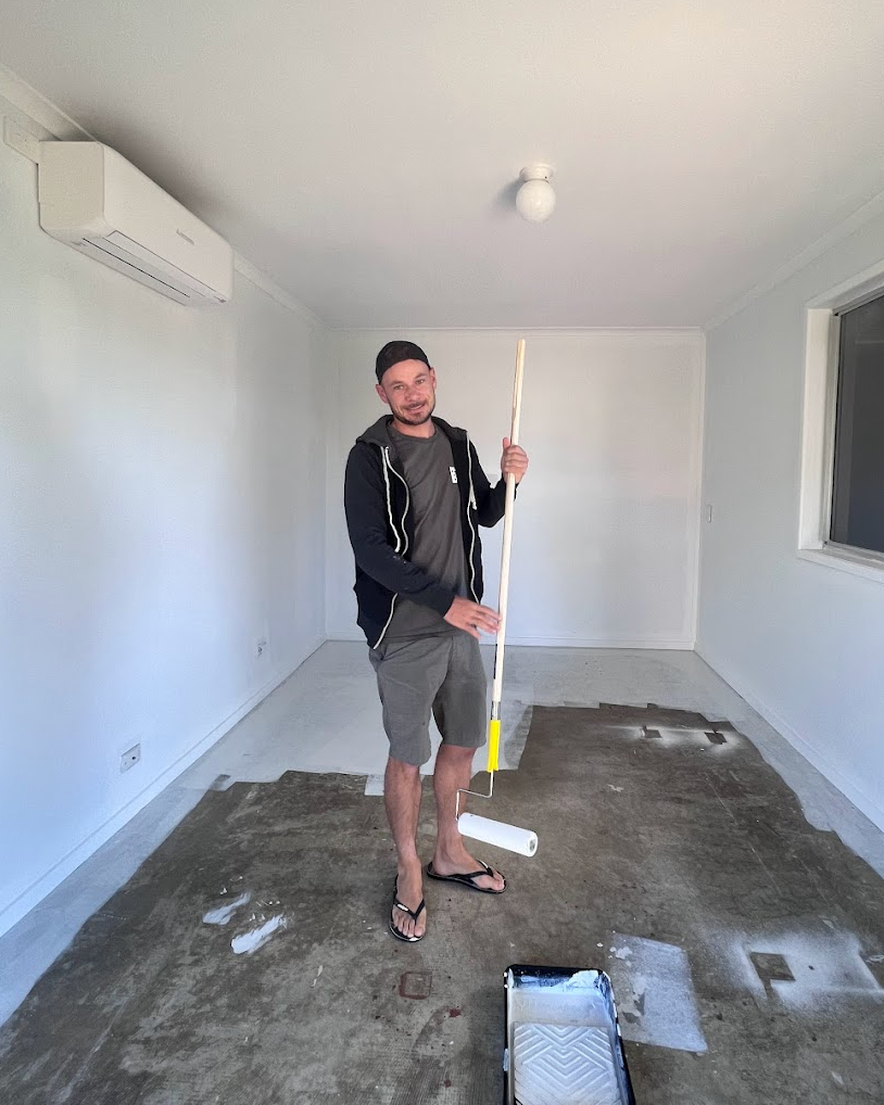 Person standing in an empty room with gray concrete floor holding a long cleaning mop next to a wall-mounted AC unit.