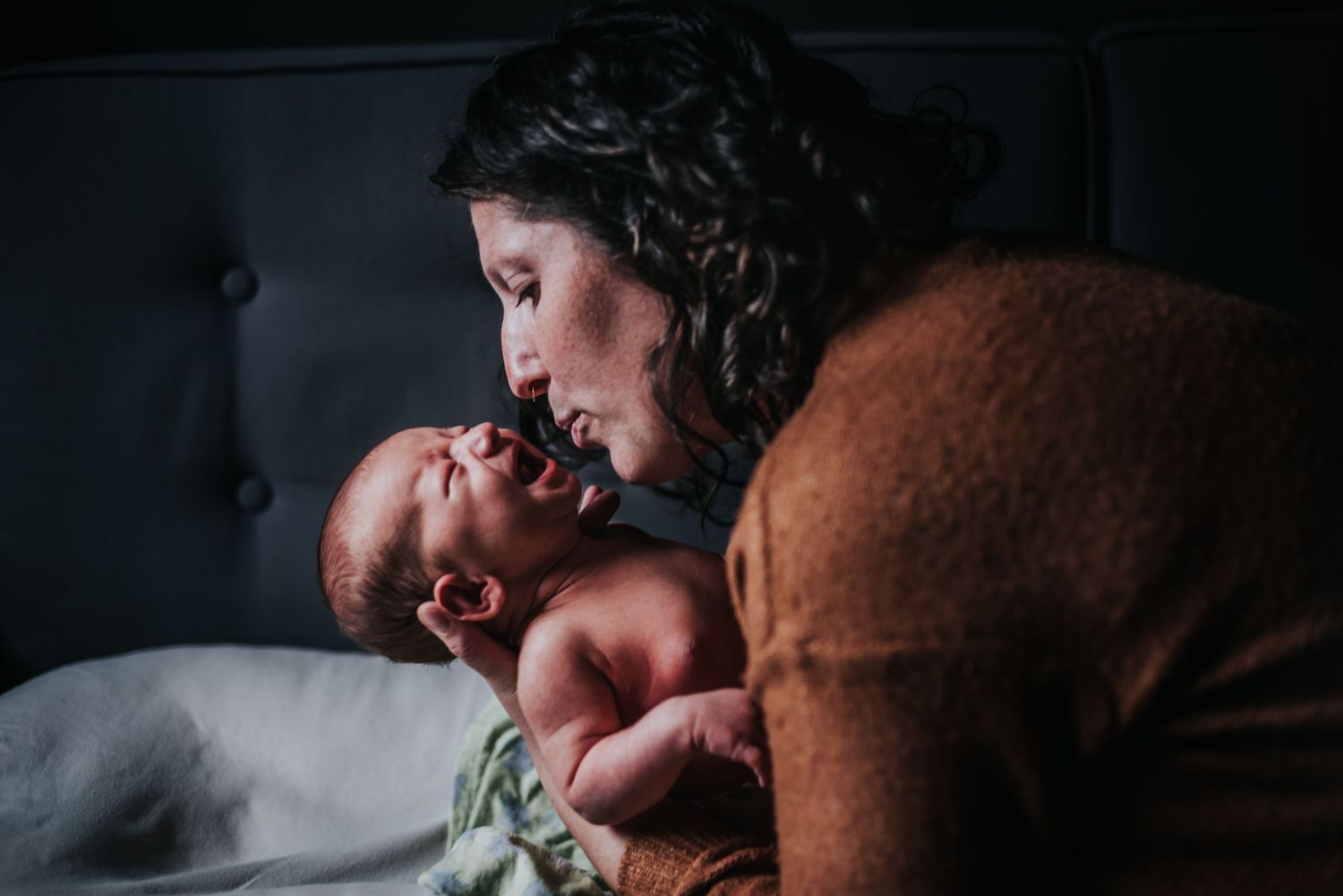Side view of parent leaning over to kiss a yawning newborn baby lying on a bed in low light.