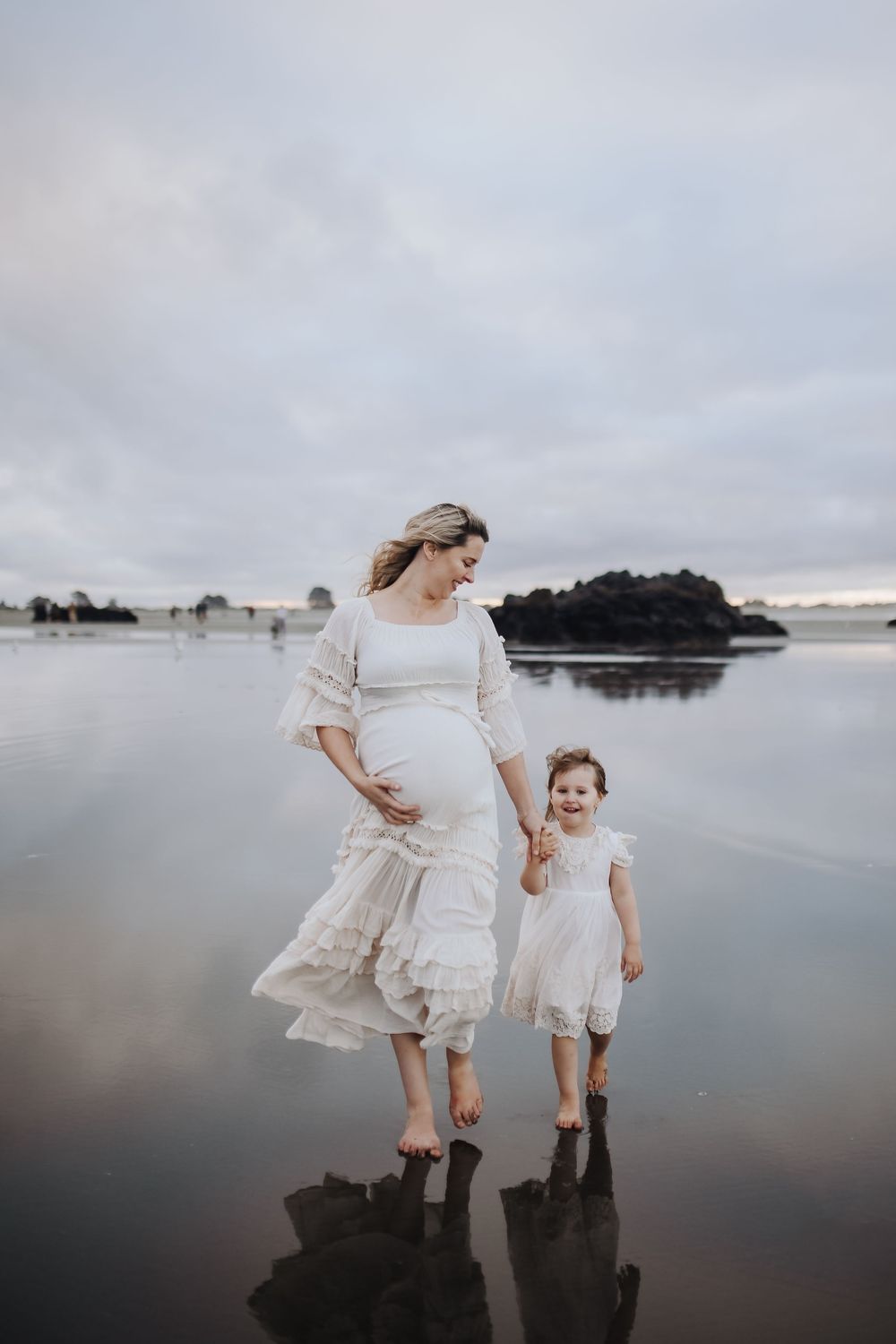Mother and child in white dresses standing on reflective beach at sunset.