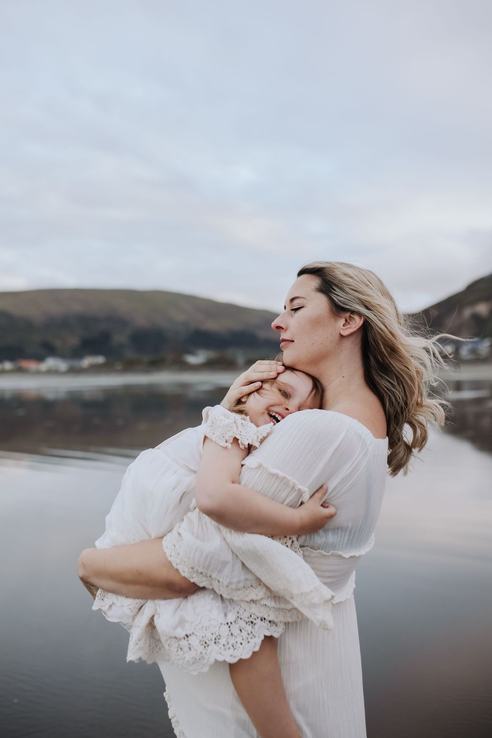 A woman holding a baby stands on a rocky shoreline with a serene lake and mountains in the background at dusk.