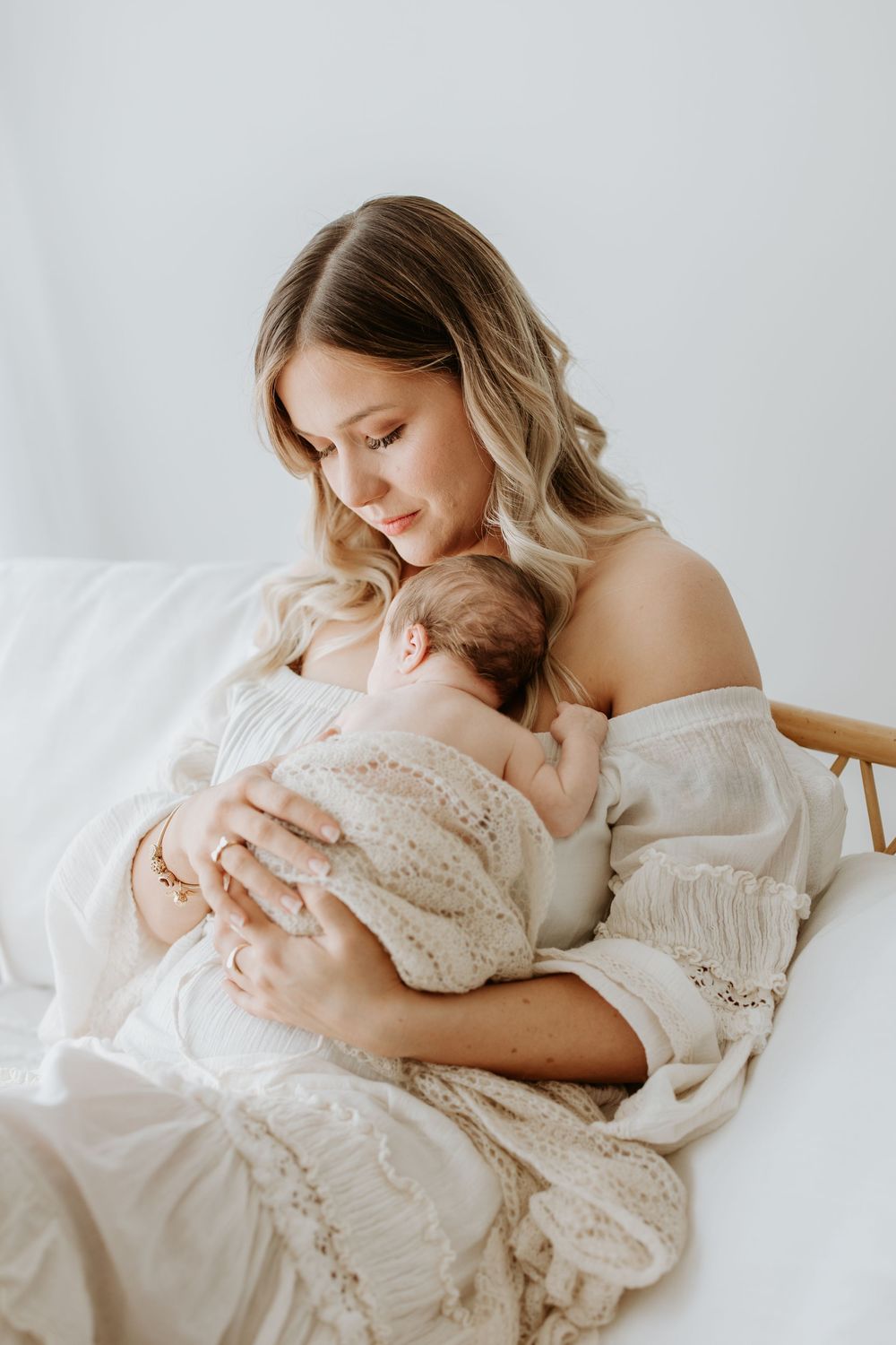 A woman in white dress cradles her sleeping newborn while sitting in a wooden chair against a light background.