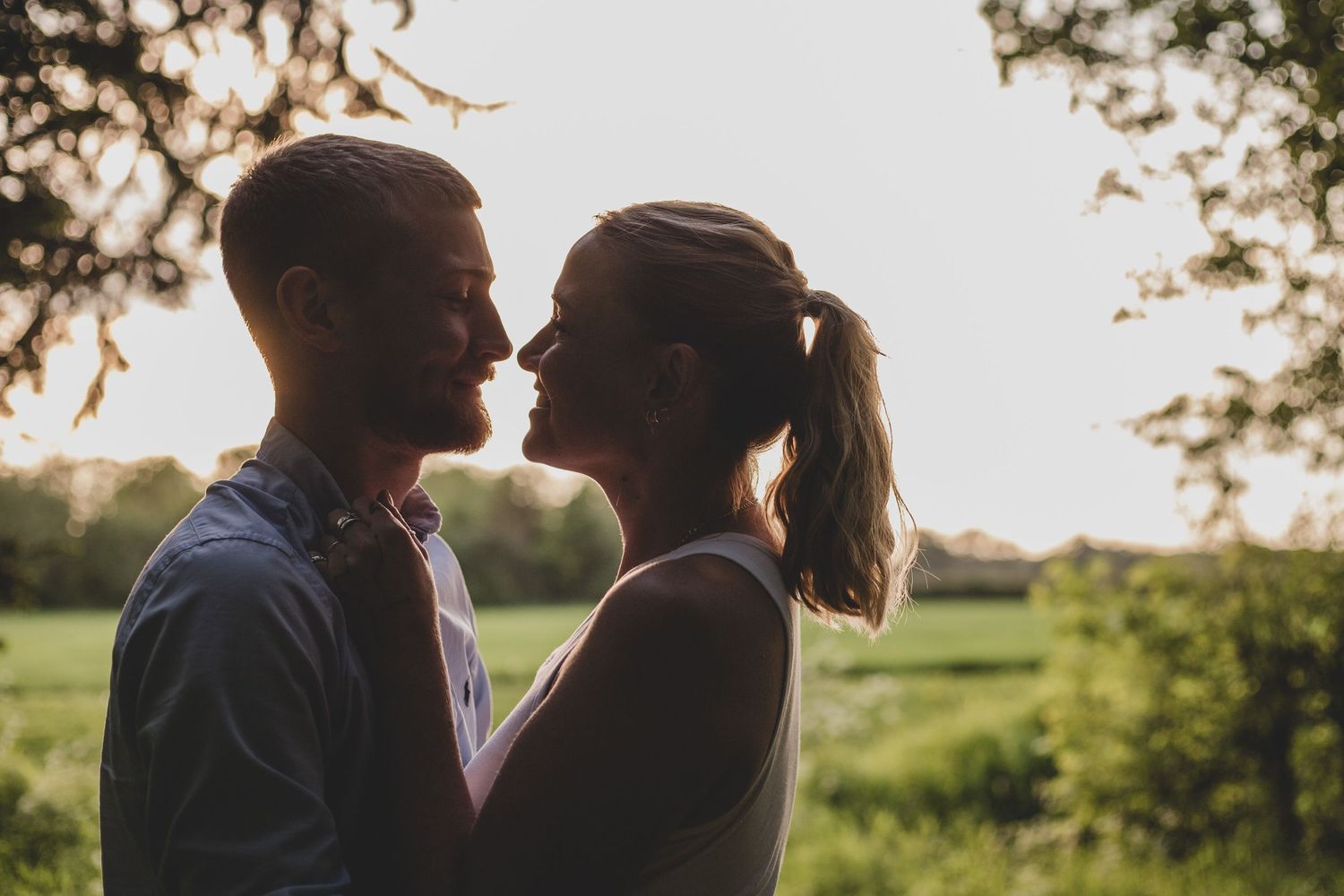 Silhouetted romantic couple in tender moment at sunset with green field and trees in background.