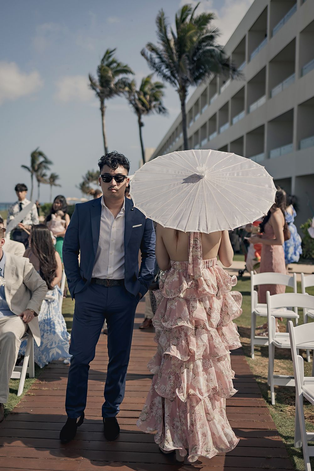 A couple walks along a wooden boardwalk in a romantic setting with white paper parasols and palm trees in the background.