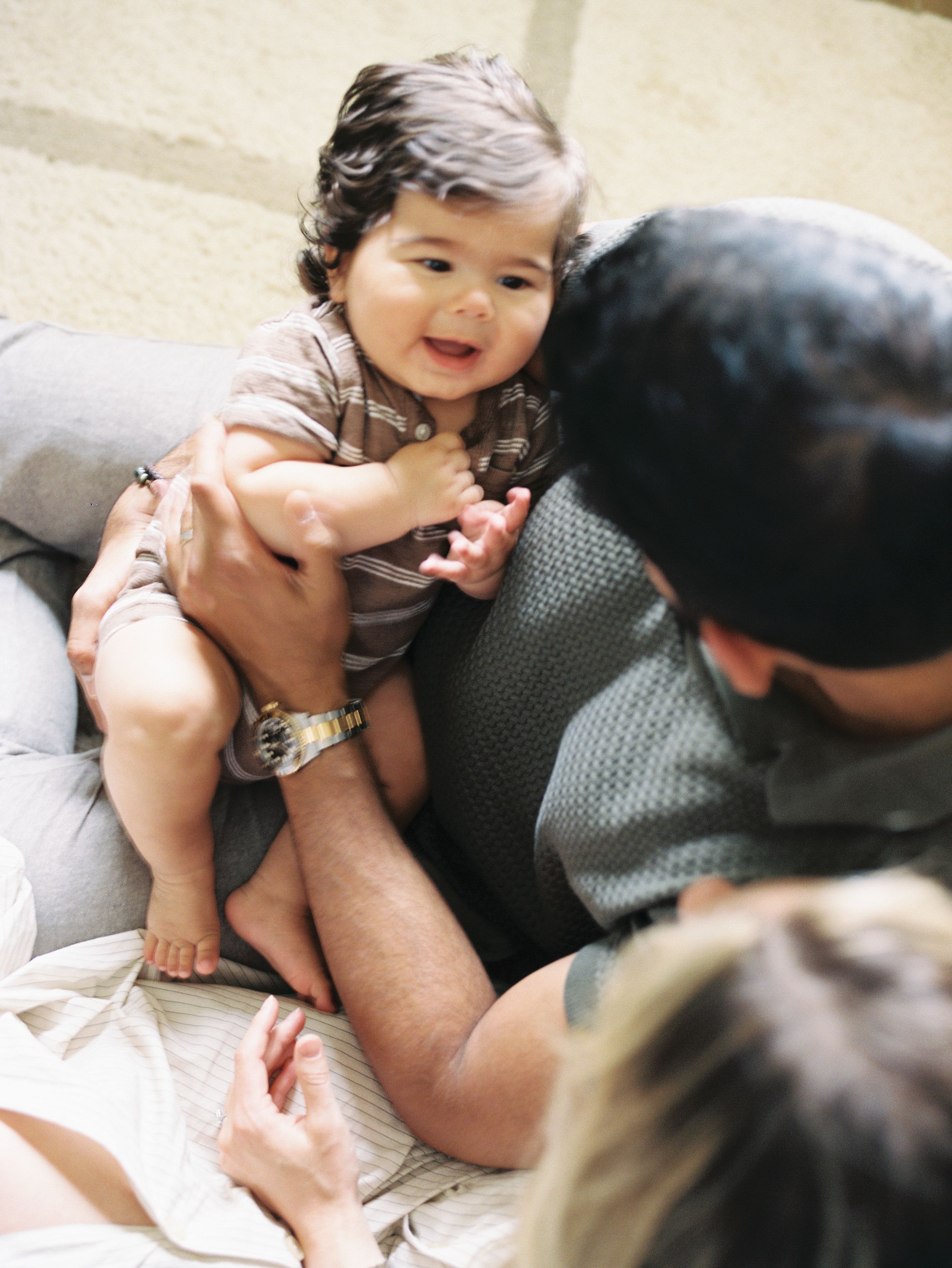 A baby wearing a striped outfit sits playfully on a gray couch while interacting with an adult.