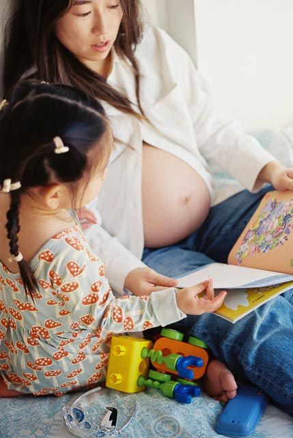 Pregnant Asian woman reading a colorful book with her young daughter surrounded by toys.